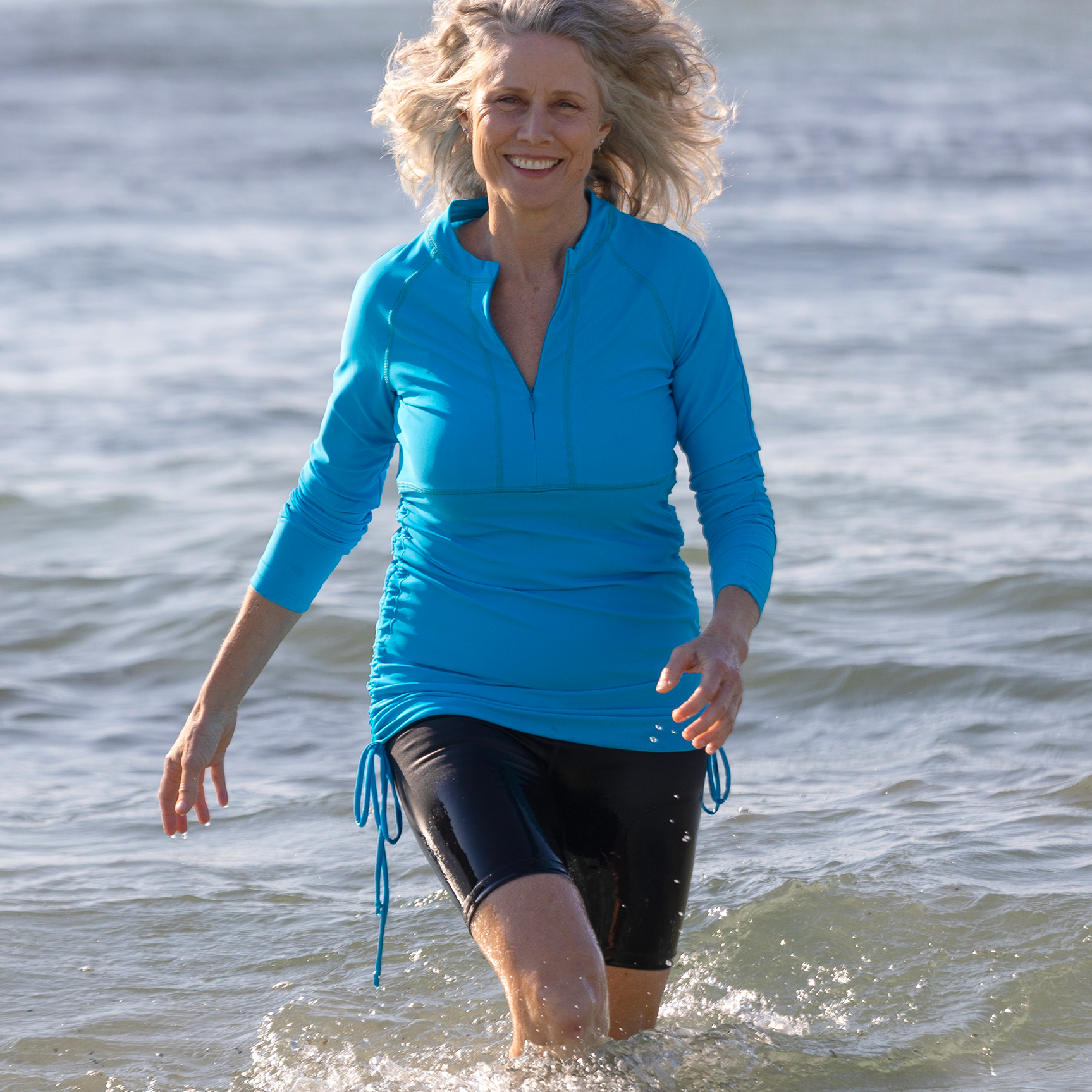 Woman in a blue long-sleeve rash top and black shorts walking in shallow water near the shore.