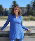 A woman smiling outdoors on a wooden boardwalk near the beach, wearing a blue long-sleeve swimdress and matching blue pants with a subtle tonal floral pattern.