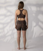 A woman stands with her back to the camera wearing a  brown and black leopard-print aswim crop top  and high-waisted boyleg shorts, against a white wall with leaf shadows.