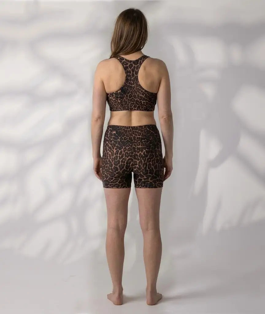 A woman stands with her back to the camera wearing a  brown and black leopard-print aswim crop top  and high-waisted boyleg shorts, against a white wall with leaf shadows.
