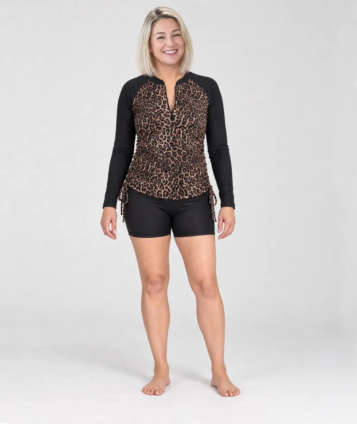 A woman wearing a long-sleeve swim top with a leopard print pattern on the front and solid black sleeves, paired with black swim shorts, against a light grey studio background.
