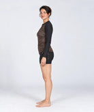 A woman stands in profile wearing a long-sleeve rash top with a leopard print pattern on the front and black sleeves, paired with black swim shorts, against a light grey studio background.
